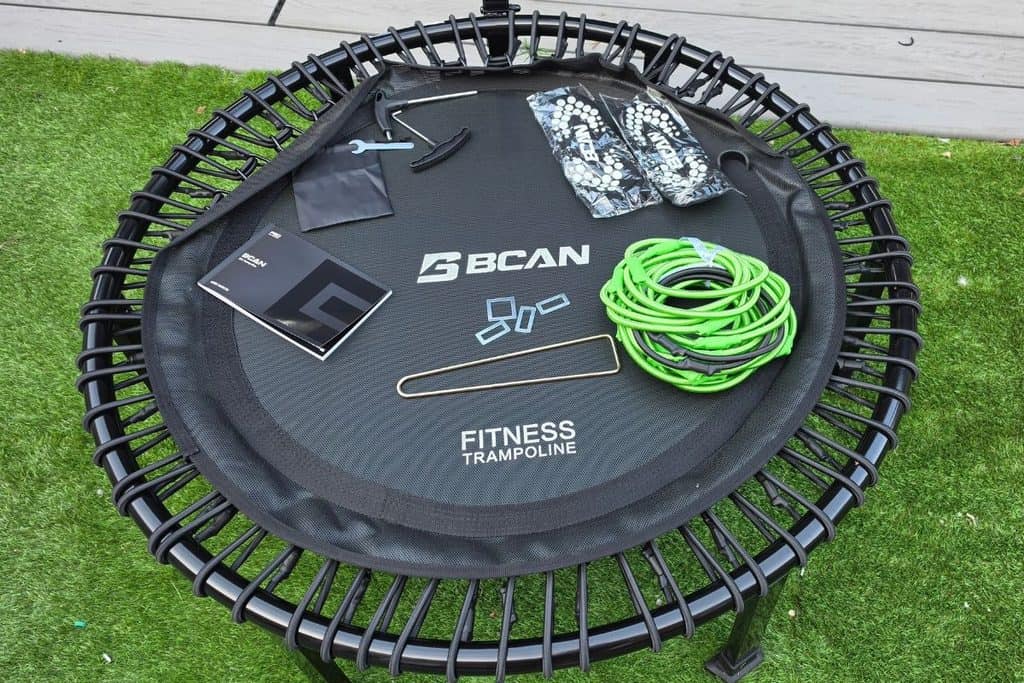 A bcan fitness trampoline and its components, including tools, springs, instruction manual, and green resistance band, are laid out on the trampoline frame on artificial grass.