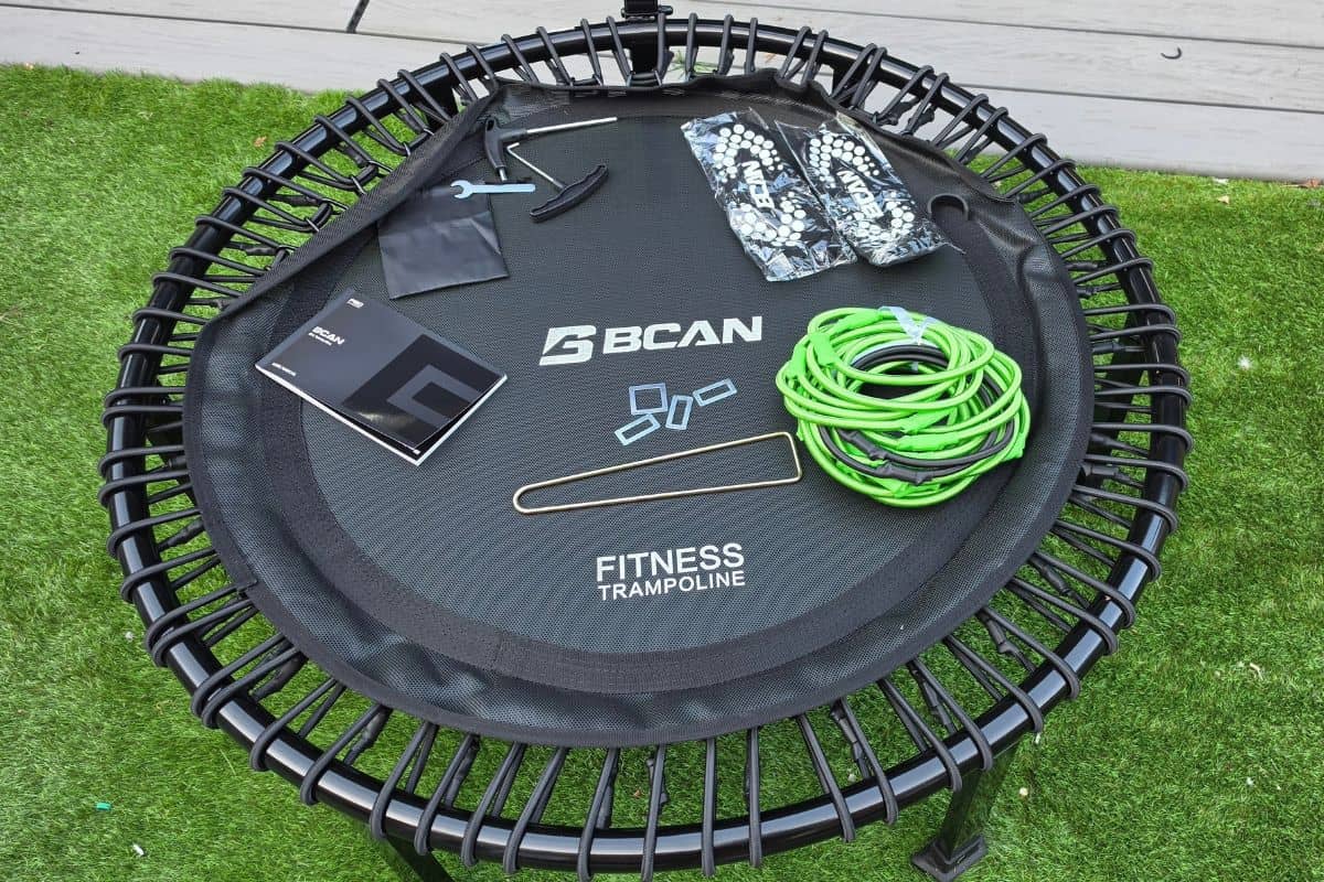 A bcan fitness trampoline and its components, including tools, springs, instruction manual, and green resistance band, are laid out on the trampoline frame on artificial grass.