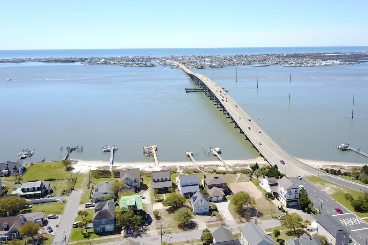 A long bridge crosses a wide body of water, connecting a residential area in the foreground to land in the distance under a clear sky.