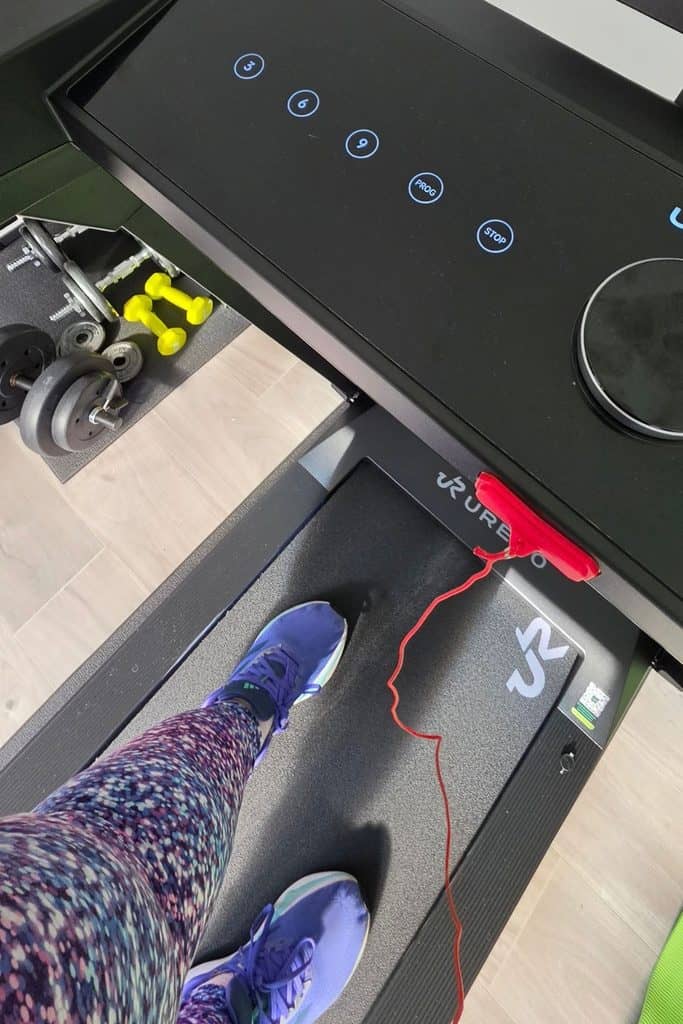 A person wearing purple trainers and patterned leggings stands on a treadmill, with dumbbells visible on the floor nearby.