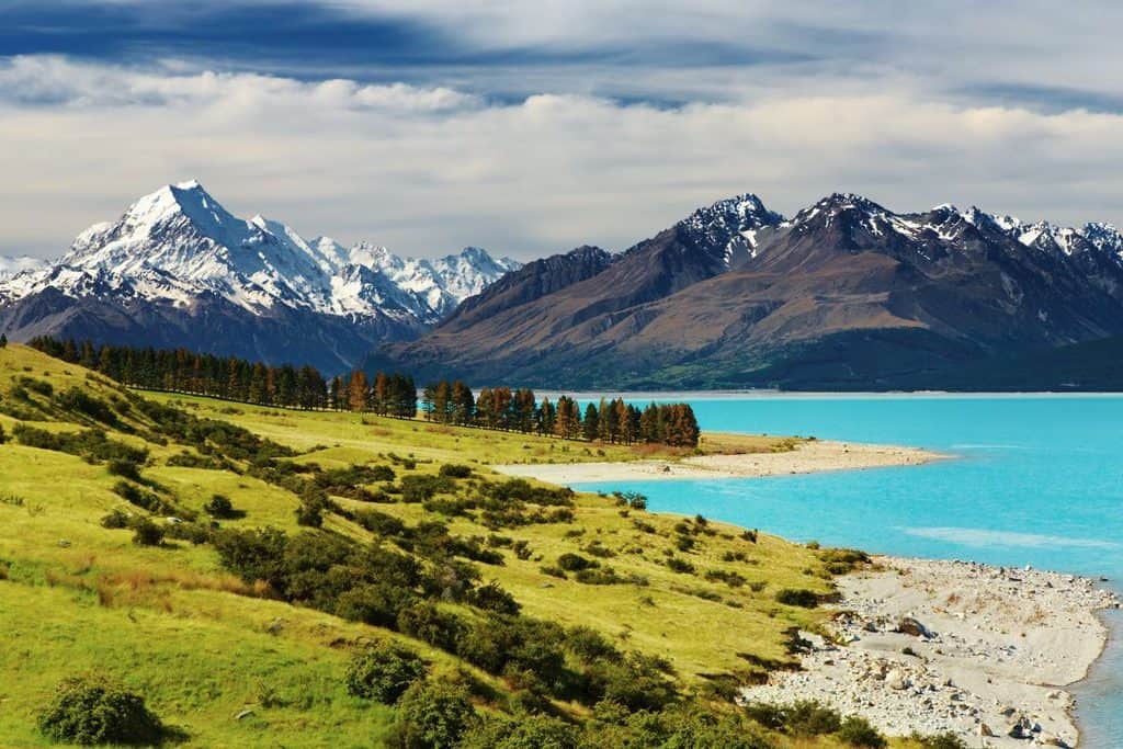 Snow-capped mountains rise behind a turquoise lake, with green hills and scattered trees in the foreground under a partly cloudy sky.