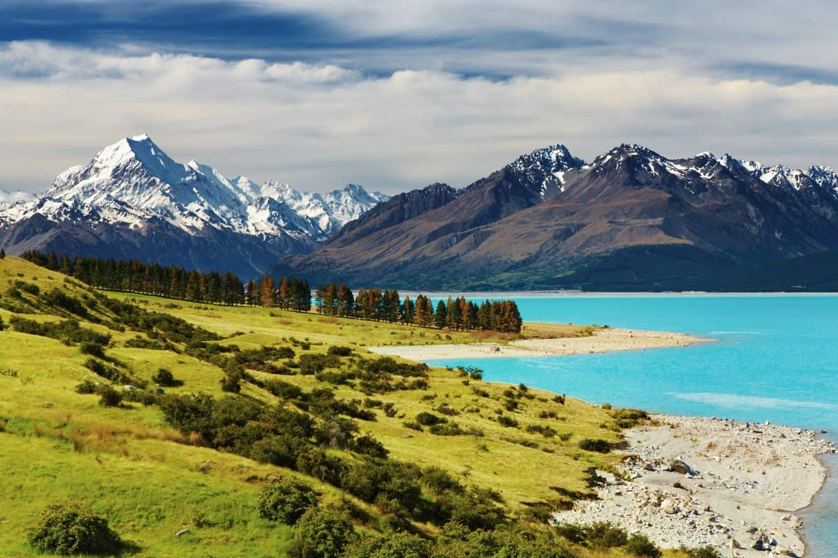 Snow-capped mountains rise behind a turquoise lake, with green hills and scattered trees in the foreground under a partly cloudy sky.