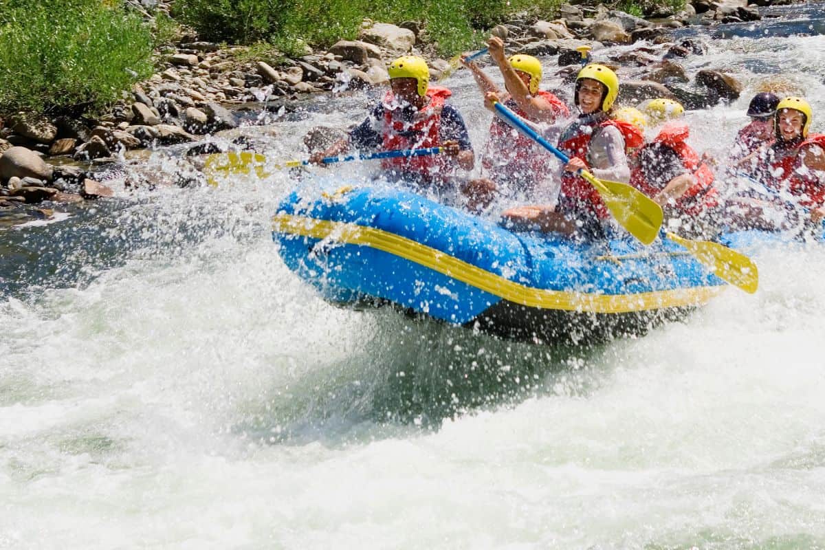 A group of people wearing helmets and life jackets paddle a blue inflatable raft through white-water rapids on a river, with water splashing around them.