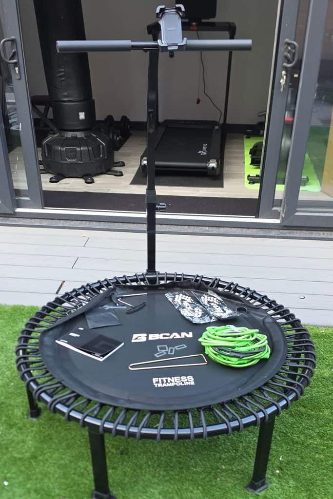 A bcan fitness trampoline with a stability handlebar, a coiled green resistance band, manual, and assembly tools on the mat.