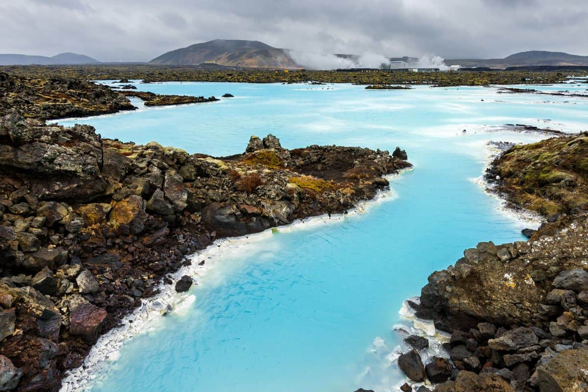 Bright blue geothermal water flows through rocky lava fields under a cloudy sky, with steam rising in the distance and mountains in the background.