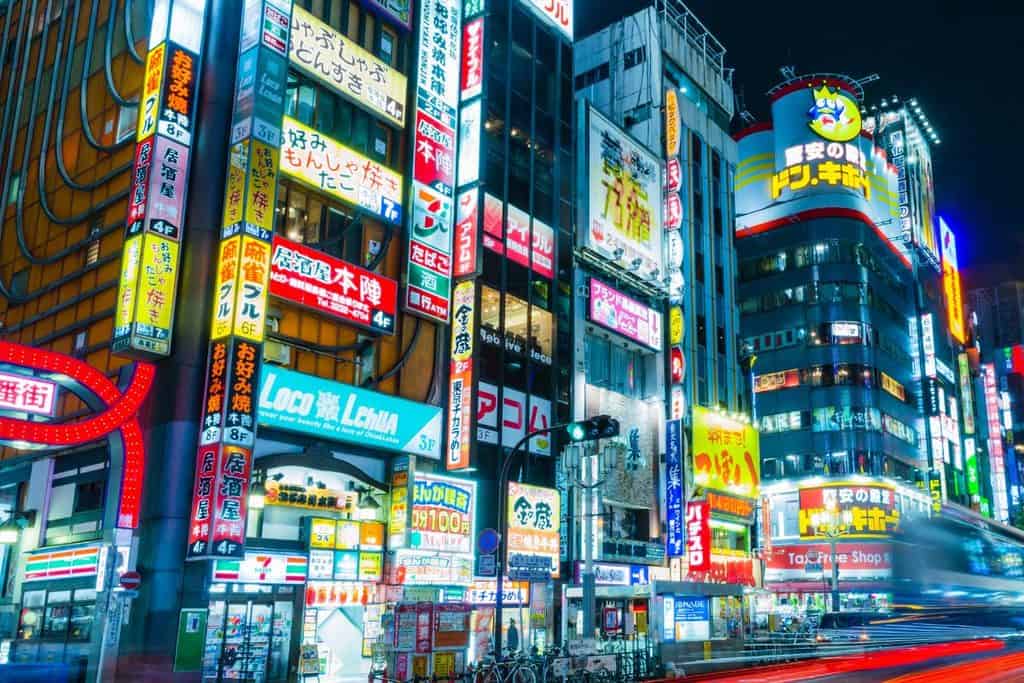 Bright neon signs and billboards cover tall buildings along a busy city street in japan at night; some bicycles are parked in the foreground.