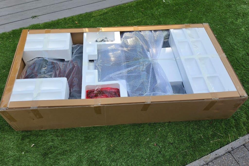 An open cardboard shipping box sitting on grass, containing packaged components for a revo brand product surrounded by protective foam and plastic wrap.